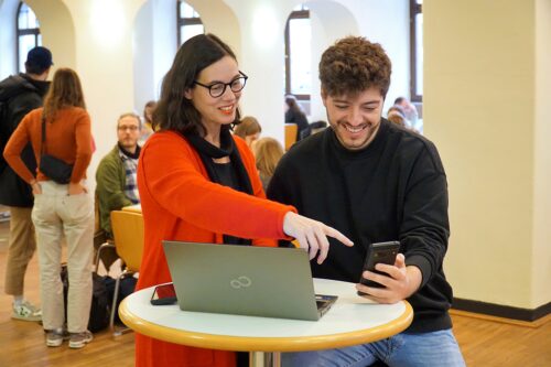 Zu sehen sind Birgitta Hohenester (links) und Max Wetterauer (rechts) in der Mensa an einem Laptop sitzend, schauen auf ein Handy.