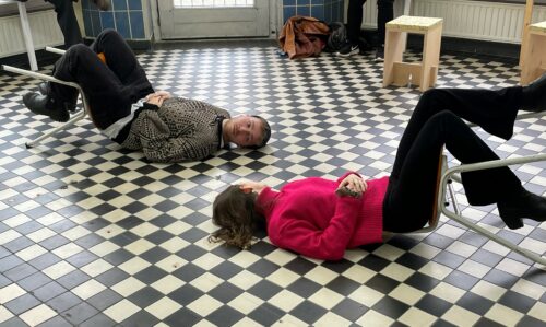 Zu sehen ist eine Kunstinstallation. Zwei Menschen liegen auf einem gekachelten Boden und blicken sich an.