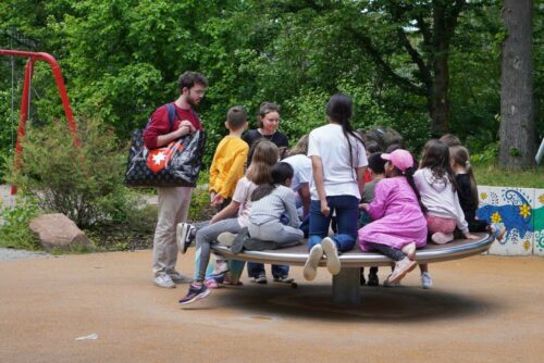 Zu sehen sind viele Kinder und 2 Studierende auf einem Balance-Gerät auf dem Spielplatz