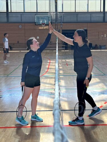 Zu sehen sind zwei Badmintonspielerinnen, die sich am Netz abklatschen