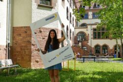 Zu sehen ist Sara Bechtel vor dem Altbau im Innenhof, wie sie durch einen Fotorahmen schaut